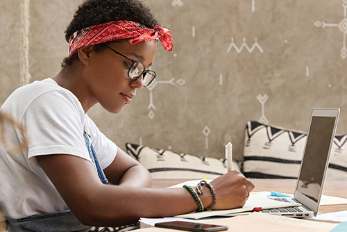 A young Black woman with glasses and a red bandana tied around her hair writes on a paper while looking at a tablet screen